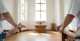 Schlichter Kirchenraum in warmem Licht. Einige Menschen sind ruhig versammelt, sichtbar sind vor allem Hände und Rückenansichten. Die Szene wirkt offen, gemeinschaftlich und nachdenklich.