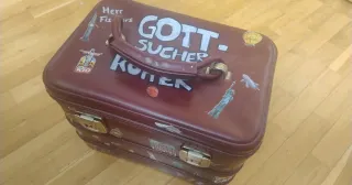 Geöffneter Koffer auf einem Tisch mit symbolischen Gegenständen wie Stein, Feder, Lupe, Tuch und Naturfundstücken als Bild für die Vorbereitung eines Gott-Sucher-Koffers für den Religionsunterricht.