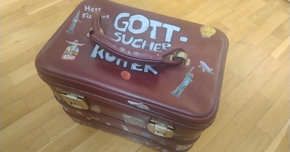 Geöffneter Koffer auf einem Tisch mit symbolischen Gegenständen wie Stein, Feder, Lupe, Tuch und Naturfundstücken als Bild für die Vorbereitung eines Gott-Sucher-Koffers für den Religionsunterricht.
